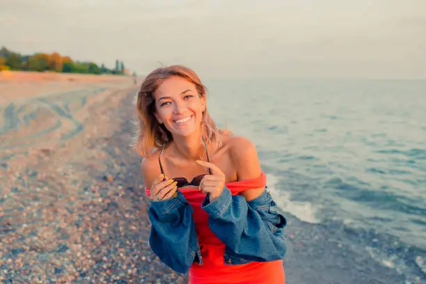 Photo of Outdoor fashion portrait of stylish girl wearing trendy sunglasses and jeans jacket on the beach. Hands up, retro filter, flecks of sunlight