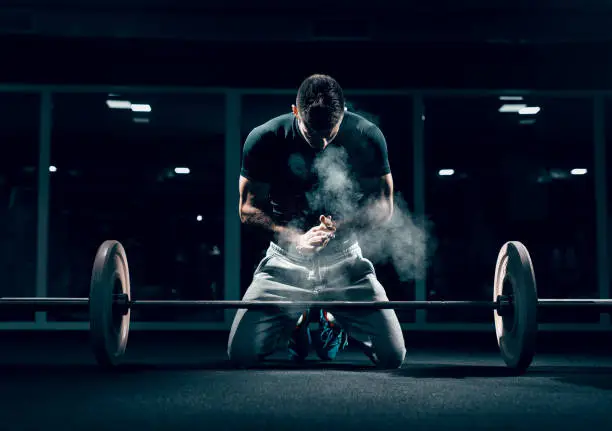 Photo of Caucasian muscular man kneeling and clapping hands. In front of him barbell, in background mirror. Gym interior, chalk all around.
