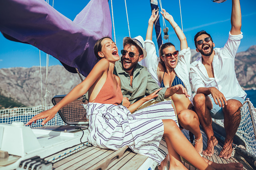 Group of friends, three women and two men, jumping  in swimwear from boat deck of a sailboat in the sea, view from the side with island and sky in the background