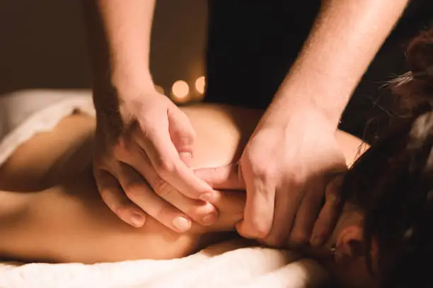 Photo of Men's hands make a therapeutic neck massage for a girl lying on a massage couch in a massage spa with dark lighting. Close-up. Dark Key
