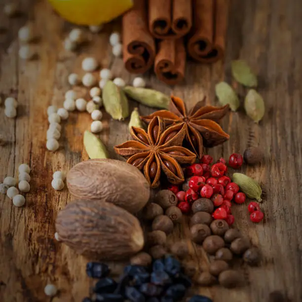 Photo of Aniseed (anise seed), barberry, cardamom, red pepper, white pepper, nutmegs, and cinnamon sticks on the wooden surface (background) of the kitchen table among the spices