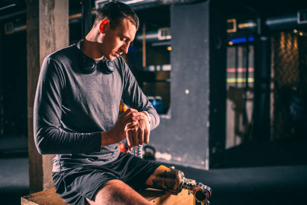 istock Young boy athlete with a prosthetic leg adjusting a fitness tracker watch while sitting on a  box in a gym gym arena 1126547271