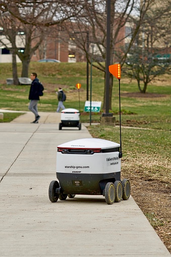 Fairfax, Virginia, USA - January 29, 2019:Fairfax, Virginia, USA - January 29, 2019: Two newly operational delivery robots travel  enroute to customers on George Mason University's main campus as students walk by. The autonomous robots are operated by Starship Technologies and use artificial intelligence technologies to securely deliver food, drinks, and snacks to users anywhere on campus via a mobile app.