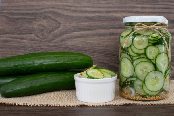 Green small cucumbers on wooden board for cooking in front of glasses with whole cucumbers, with mustard seeds and onions, with no dark wood background and wooden spoon. stock photo