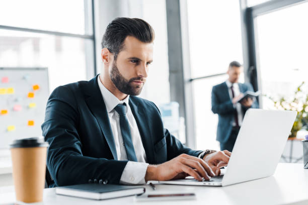 istock focused businessman typing on laptop with coworker on background 1126564517