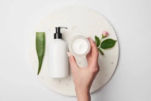 Photo of partial view of female hand near cream container with cosmetic dispensers, aloe vera leaf and rose flower on white surface