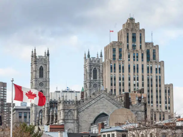 Photo of Skyline of Old Montreal, with Notre Dame Basilica in front, a vintage stone Skyscraper in background & a Canadian flag waiving. The basilica is the main cathedral of Montreal, Quebec, Canada, & a touristic landmark