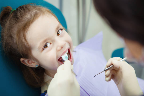 Happy child in dentist chair with napkin on chest Happy child in comfortable dentist chair with paper napkin on chest and doctor in rubber gloves who checks mouth with sharp metal instrument and magnifying glass. pediatric dentistry stock pictures, royalty-free photos & images