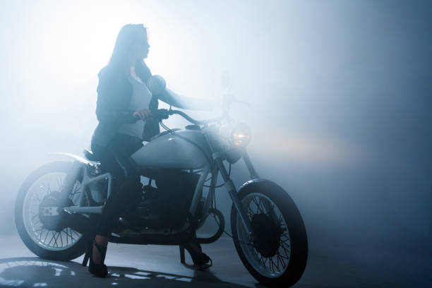 Slender young woman with long straight hair is sitting on restored retro motorcycle in a half turn to the camera. Studio shooting with a smoke, silhouette backlit