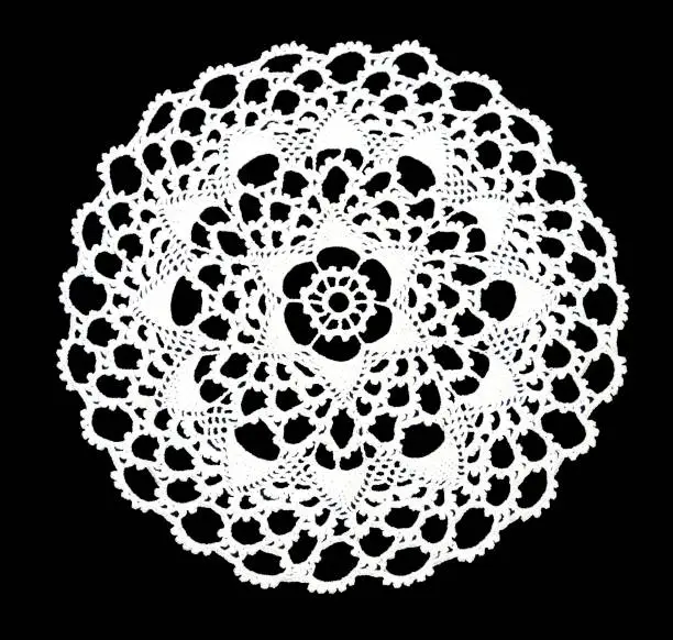 Isolated crocheted laced white doily with many arches and picos at the egde on a black background. Round decorative cotton doily. Top view