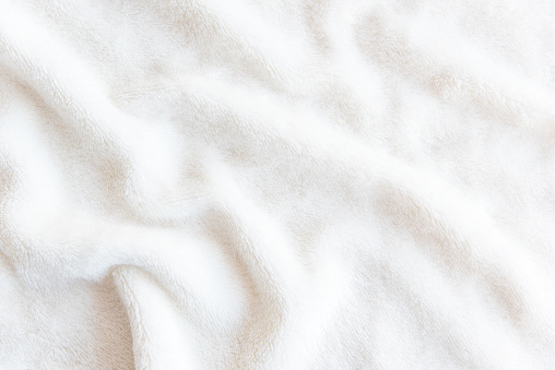 White delicate soft  background of plush fabric. Texture of beige soft fleecy blanket textile with twisted folds.