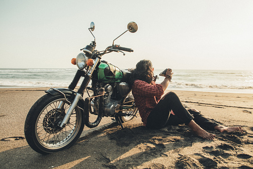 One man, handsome man sitting on sand by his motorcycle on the beach, taking pictures with instant camera.