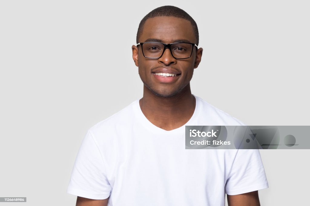 Smiling african millennial man looking at camera isolated on background Smiling african american millennial casual man wearing white t-shirt and glasses looking at camera isolated on blank grey studio background, charming young black single male guy posing for portrait Men Stock Photo
