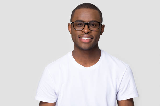 Smiling african millennial man looking at camera isolated on background Smiling african american millennial casual man wearing white t-shirt and glasses looking at camera isolated on blank grey studio background, charming young black single male guy posing for portrait black nerd stock pictures, royalty-free photos & images