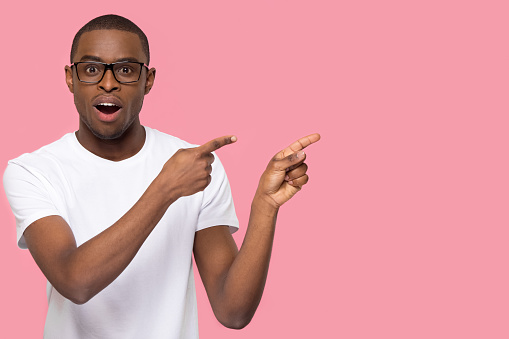 Surprised black man pointing fingers at copy space looking at camera, amazed african american guy feeling astonished excited advertising product or service isolated on pink blank studio background