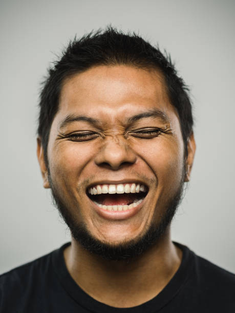 Real malaysian young man with very excited expression and eyes closed Close up portrait of asian young man with very excited expression against white gray background. Vertical shot of malaysian real people laughing and shouting in studio with black hair and beard. Photography from a DSLR camera. Sharp focus on eyes. man laughing hysterical stock pictures, royalty-free photos & images