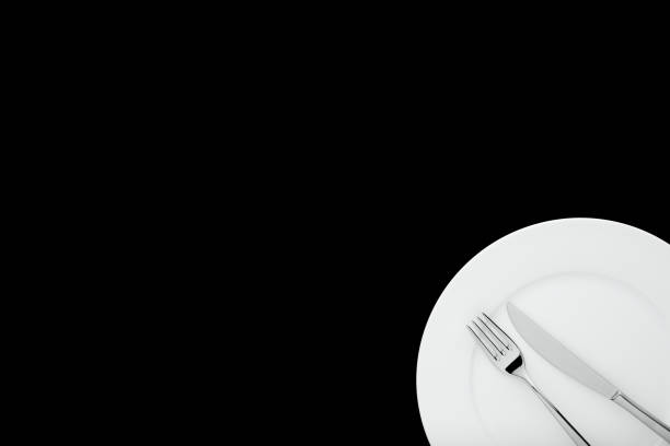 Fork and knife on white round ceramic plate on isolated black background with copy space. Top down view.
