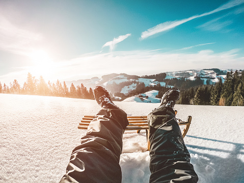 Pov view of young man looking the sunset on snow high mountains with vintage wood sledding - Legs view of travel influencer creating content - Winter vacation concept - Focus on his feet