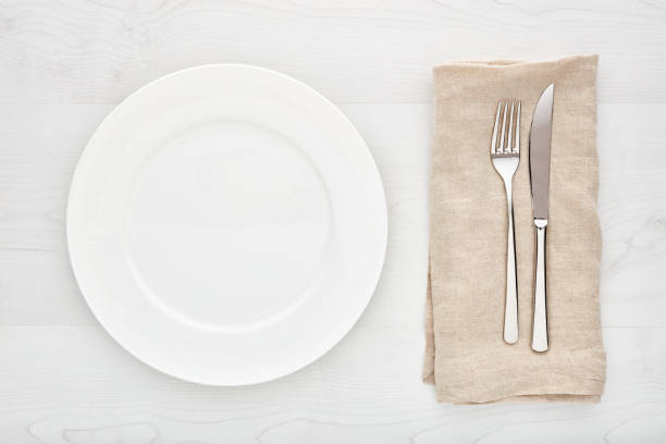 istock Empty white round ceramic plate, fork, knife and napkin on white wooden table. Top down view. 1126668212