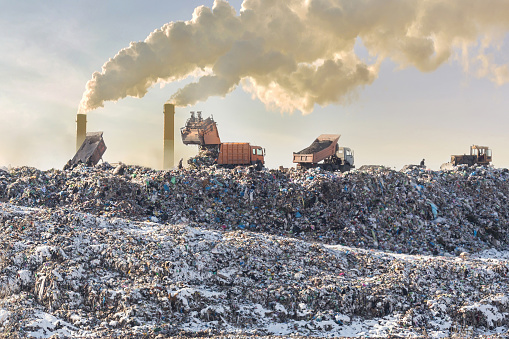 Dump trucks unloading garbage over vast landfill. Smoking industrial stacks on background. Environmental pollution. Outdated method of waste disposal. Survival of times past.