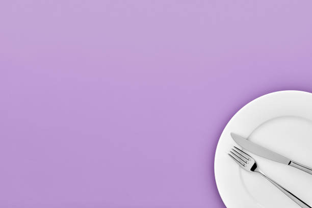 istock Fork and knife on white round ceramic plate on light purple background with copy space. Top down view. 1126669042