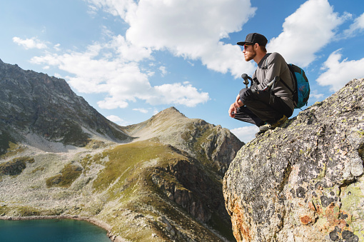 A bearded hipster tourist in sunglasses with a backpack sits on the edge of a cliff high in the mountains near the mountain lake.