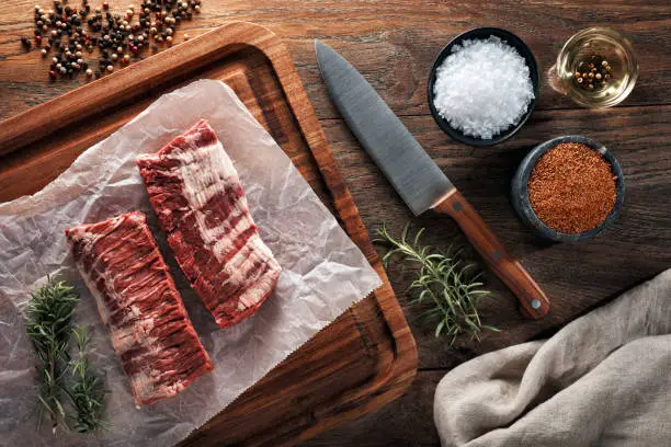 Raw calf skirt steak meat on white cooking paper and wooden cutting board. Decorated with herbs, spices and chef's knife. Overhead view.