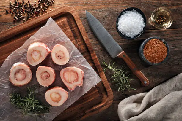 Raw calf bones on white cooking paper and wooden cutting board. Decorated with herbs, spices, chef's knife and napkin. Overhead view.