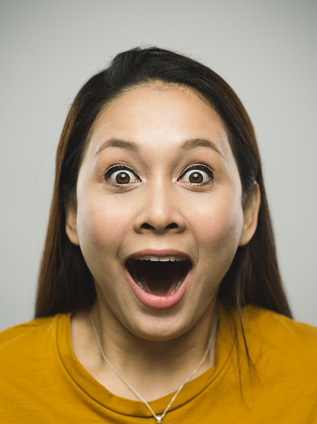 Close up portrait of asian young woman with surprised expression against white gray background. Vertical shot of malaysian real people laughing and surprised in studio with long brown hair. Photography from a DSLR camera. Sharp focus on eyes.