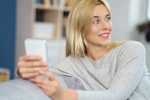 Young blond woman sitting on a sofa with her mobile in her hands watching something to the side with a smile