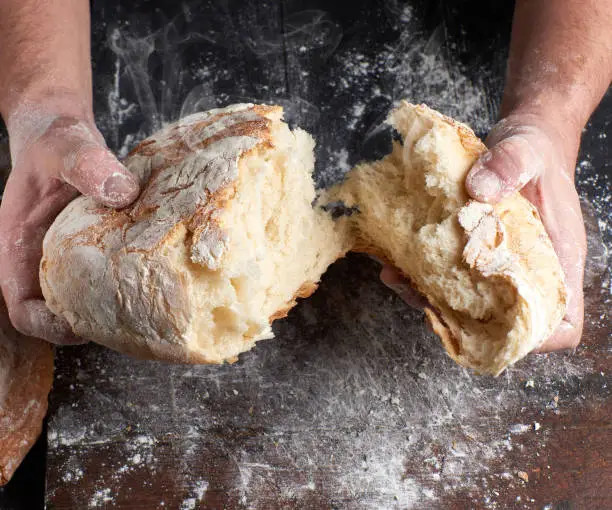 male hands breaking open baked bread in half over black wooden table