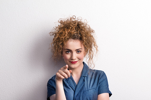 Young beautiful happy woman standing in studio, looking at camera and finger pointing at you.