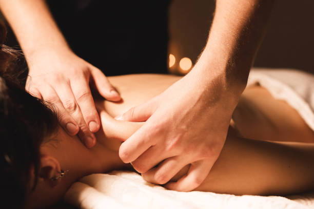 Men's hands make a therapeutic neck massage for a girl lying on a massage couch in a massage spa with dark lighting. Close-up. Dark Key stock photo