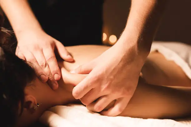 Photo of Men's hands make a therapeutic neck massage for a girl lying on a massage couch in a massage spa with dark lighting. Close-up. Dark Key