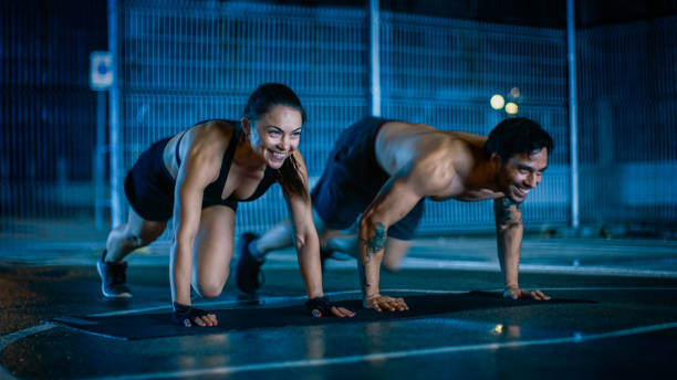 sorrindo feliz atlética fitness casal fazendo montanha alpinista exercícios. treino é feito em uma quadra de basquete ao ar livre cercado. à noite depois da chuva em uma área residencial do bairro. - people recreational pursuit exercising physical activity - fotografias e filmes do acervo