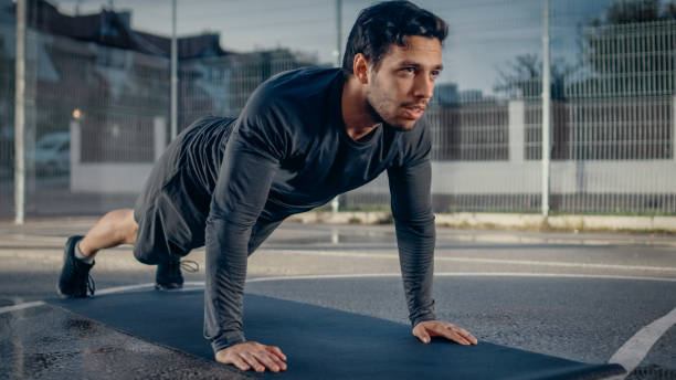 Strong Muscular Fit Young Man in Sport Outfit Doing Push Up Exercises. He is Doing a Workout in a Fenced Outdoor Basketball Court. Evening After Rain in a Residential Neighborhood Area.