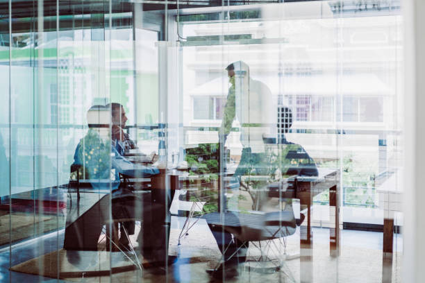 Business colleagues in meeting at office Business colleagues in meeting at office. Male and female professionals are planning strategy in brightly lit workplace. They are seen through glass wall with reflections. corporate boardroom stock pictures, royalty-free photos & images