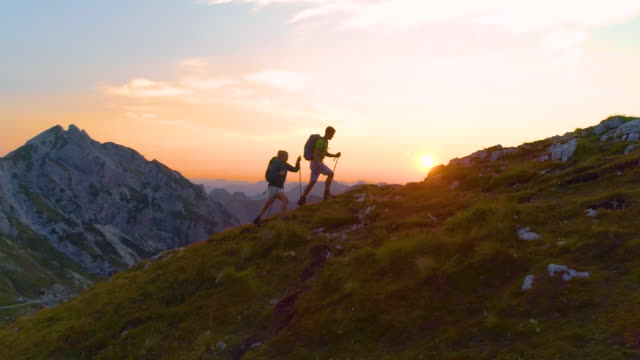 AERIAL: Active young tourists hiking up a grassy mountain in the Alps at sunset.