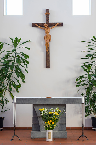 The Altar of a catholic church, in a clean design with some green plants and a bouquet of yellow flowers in front of the altar table. There is a wooden crucifix on the wall.