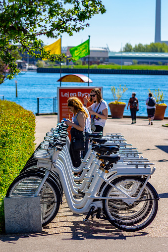 City bikes (Bycyklen) in docking stations in Copenhagen, Denmark. The bikes are available for short term hire by commuters or tourists and can be left at other docking stations around the city. There are two young women hiring bikes beside the harbour where the Little Mermaid statue is.