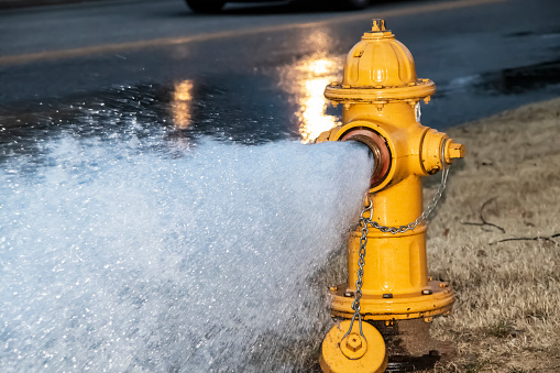 Close-up of yellow fire hydrant gushing water across a street with wet highway and tire from passing car behind