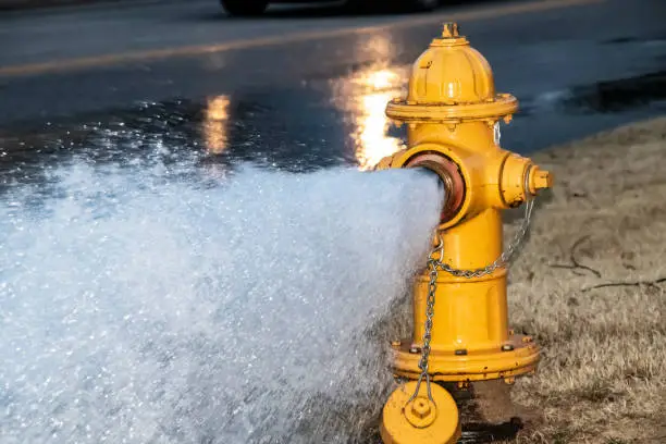 Photo of Close-up of yellow fire hydrant gushing water across a street with wet highway and tire from passing car behind