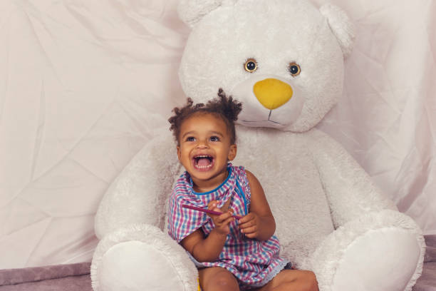 Little cute african american embarrassed baby girl with broad smile. Lovely kid looking happy and wearing bright plaid dress, lying on a big soft teddy bear