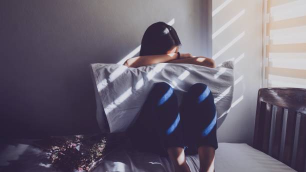 asiatische frauen sitzen die knie im bett umarmt. traurig, enttäuscht, verliebt in die dunklen schlafzimmer und das sonnenlicht aus dem fenster durch die jalousien. vintage-ton. - depression stock-fotos und bilder