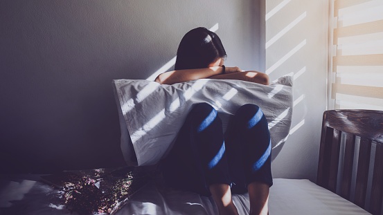 Las mujeres asiáticas son sentada abrazando sus rodillas en la cama. Sentirse triste, decepcionada en el amor en el dormitorio oscuro y la luz del sol desde la ventana a través de las persianas. Tono vintage. photo