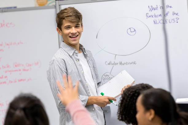 Teenage boy takes questions during high school presentation A cheerful male high school student stands before a group of classmates during a presentation and listens as an unrecognizable classmate raises a hand and asks a question. hand raised classroom student high school student stock pictures, royalty-free photos & images
