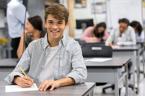 A confident teenage boy sits at a desk in his high school classroom and pauses from taking an exam to smile for the camera.