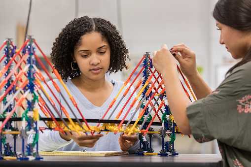 Two teen girls sit at a table in their high school classroom and build a bridge using a model kit.  One girl looks down in concentration.