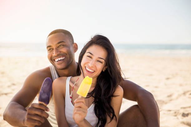 A photo of happy couple holding popsicles while sitting at beach. Cheerful man and woman enjoying summer vacation. Smiling male and female are relaxing on sunny day.