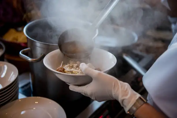Photo of Cooking Noodle soup : Hand of Thai Chef pour boiling water soy soup to ramen, noodles with fish ball in bowl at restaurant shop, Thai street food market, making noodle in Traditional Asia Food.